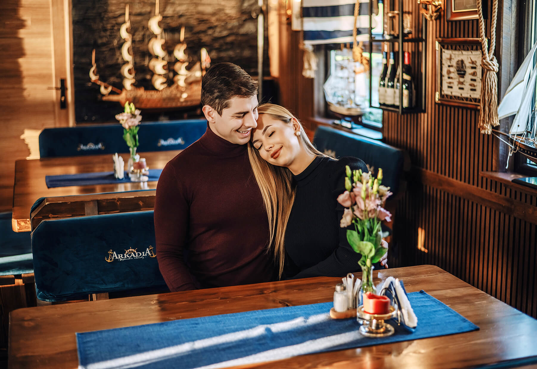 Para siedzi blisko siebie w przytulnej restauracji na Wiśle w Krakowie. Kobieta opiera głowę na ramieniu mężczyzny, oboje się uśmiechają. Kwiaty i nakrycia zdobią drewniany stół przed nimi.