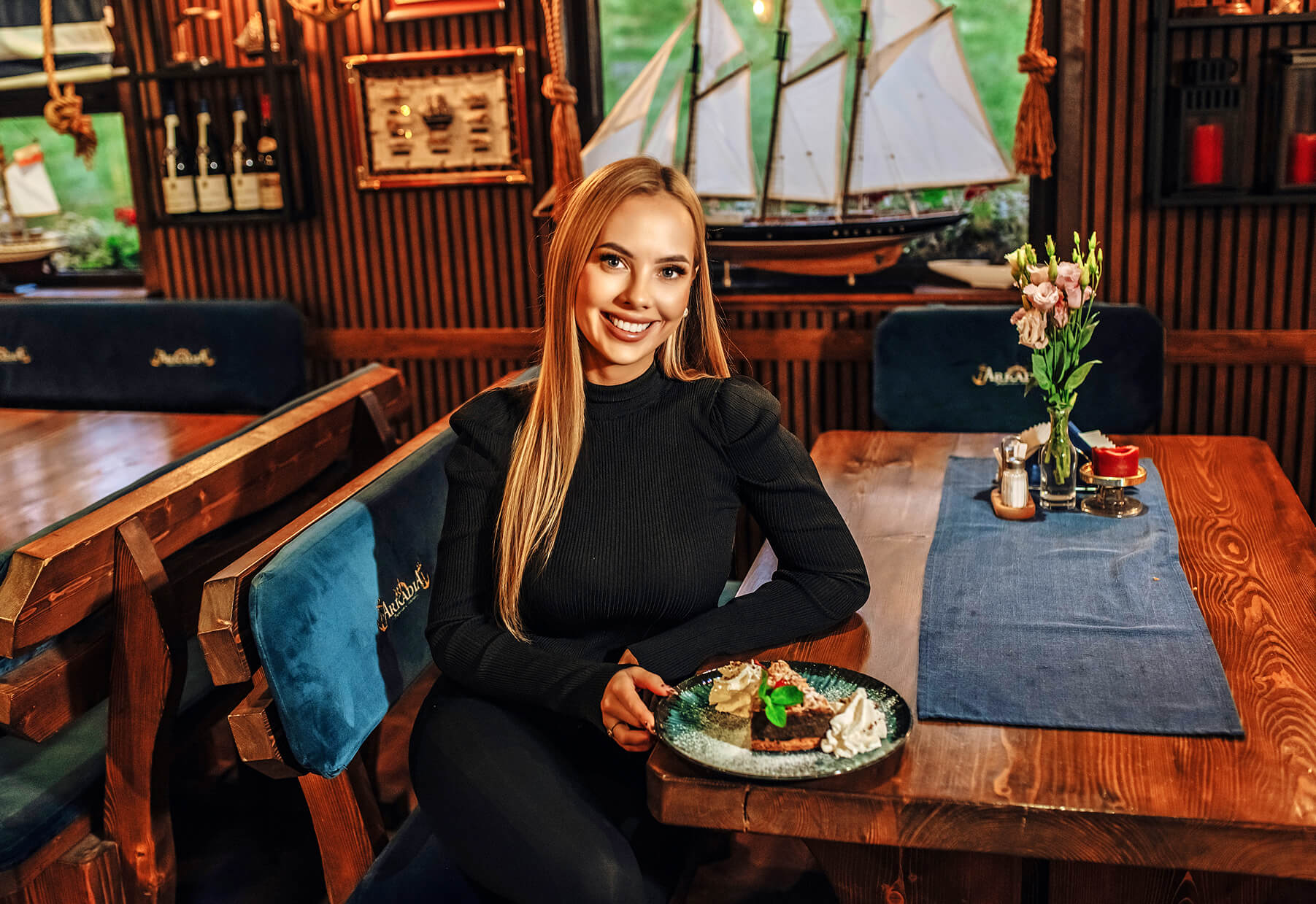 Uśmiechnięta kobieta z długimi blond włosami siedzi przy drewnianym stole w restauracji na Wiśle w Krakowie, trzymając talerz z deserem. Modele statków, kwiaty i butelki wina zdobią przytulne, ciepło oświetlone tło o tematyce żeglarskiej.